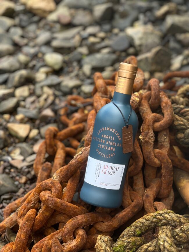 Ardnamurchain whisky glass bottle made by Verallia on a rock beach surrounded by rusting chains